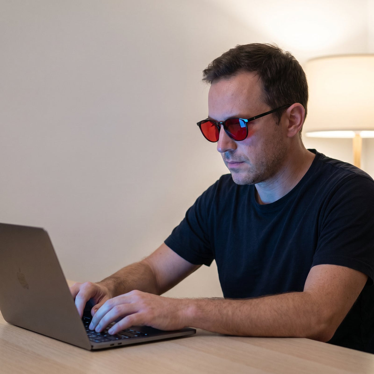 A man working at a laptop in the evening wearing Hueban SR Prism glasses with black round frames and red lenses
